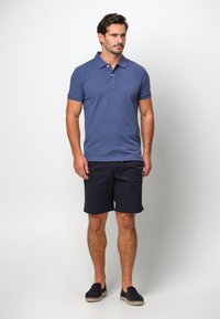 Camisa polo azul marino con cuello y mangas cortas, combinada con pantalones cortos oscuros. Tejido ligero y diseño sencillo. Zapatillas casuales sin cordones.