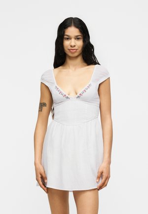 Tânără cu păr lung și închis la culoare, purtând o rochie albă scurtă cu broderie florală la decolteu și un tatuaj pe brațul stâng.