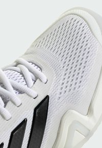 Bílá sportovní obuv s prodyšným síťovaným svrškem, plochými bílými tkaničkami a černými pruhy jako akcentem. Texturovaná podrážka s výrazným tvarem.