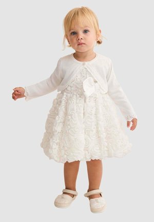 Bambina con capelli biondi che indossa un vestito bianco con motivo floreale e texture, un cardigan abbinato e scarpe beige a cinturino, in piedi davanti a uno sfondo neutro.