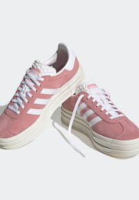 Un par de zapatillas Adidas de ante rosa con rayas blancas, suelas gruesas de color crudo, cordones blancos y una lengua texturizada blanca con el logo.