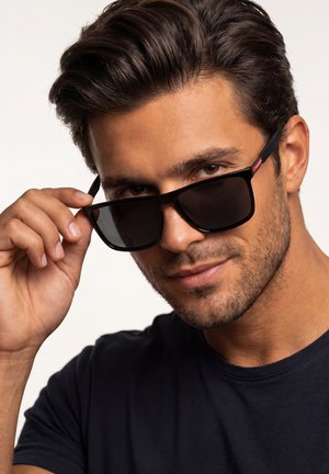 Mężczyzna z krótkimi ciemnymi włosami i zarostem opuszcza czarne okulary przeciwsłoneczne, patrząc prosto przed siebie, ubrany w ciemną koszulę na jasnym tle.