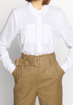 Chemise blanche à manches longues, avec une texture rayée, comportant deux poches poitrine. Associée à un pantalon beige ceinturé.