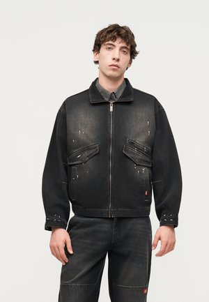 Young man wearing a black zip-up jacket and matching black pants, standing against a plain light background.