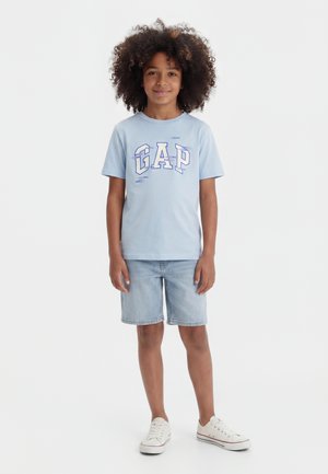 Παιδί με σγουρά μαλλιά που φοράει ανοιχτό μπλε μπλουζάκι GAP, τζιν σορτς και λευκά αθλητικά, στέκεται μπροστά σε μια λευκή απλή επιφάνεια.