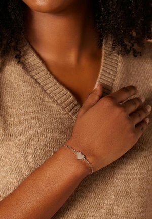 Silberne Kettenarmband mit einem kleinen Herzanhänger, vor einem beigen Pullover präsentiert, der ein glattes Metallic-Finish und ein zartes Design hervorhebt.
