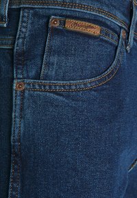 Blå jeans i denim med en brun läderetikett, kopparnitar, kontrasterande gul söm och en klassisk fem-fickdesign.