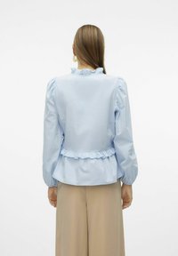 Blusa azzurra chiara con scollatura e orlo arricciati, maniche a palloncino, tessuto morbido e design a peplo. La vista posteriore mostra una vita aderente.
