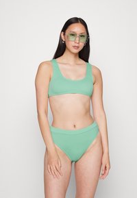 Conjunto de bikini em verde menta com duas peças, topo de decote redondo e partes de baixo de cintura alta, feito de um tecido liso e elástico. Óculos com lentes verdes.
