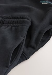 Mustad elastse vöökohaga dressipüksid küljetaskutega, volditud heledal pinnal, näidates kangastekstuur ja õmbluse detaile.