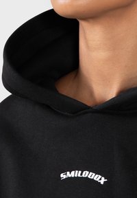 Gros plan sur une personne portant un hoodie noir avec une grande capuche et le texte blanc "SMILODOX" sur la poitrine.