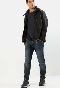 Sort, vejrbestandig jakke med hætte, lynlås og lommer; blå t-shirt, mørke jeans og blå sneakers fuldender outfittet.