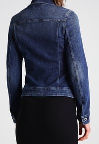 Mörkblå denimjacka, figursydd design med en något urtvättad textur, knäppta ärmslut och krage. Bakvyn visar strukturerade sömmar och fåll.