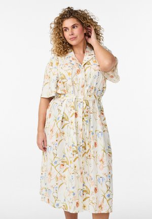 Žena s kučeravými vlasmi, ktorá má na sebe midi šaty s krátkym rukávom, gombíky a opaskom s kvetinovým vzorom na svetlom pozadí.