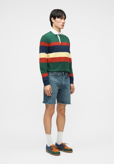 Νεαρός άνδρας που φοράει ριγέ μακρυμάνικη ράγκμπι μπλούζα, μπλε τζιν σορτς, λευκές κάλτσες και πολύχρωμα παπούτσια τύπου boat, στέκεται προφίλ.