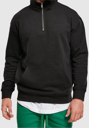 Mann trägt schwarzen Sweatshirt mit Viertelreißverschluss und Kängurutasche, darunter weißes Hemd, sowie grüne Hose mit Kordelzug vor grauem Hintergrund.