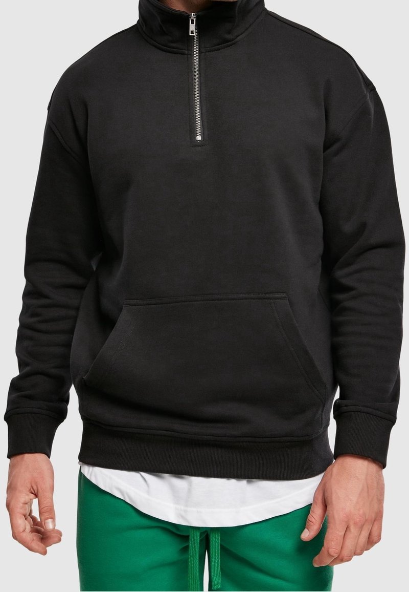 Homme portant un sweat-shirt noir à demi-fermeture éclair avec poche kangourou, chemise blanche en dessous, et pantalon vert à cordon de serrage sur fond gris.