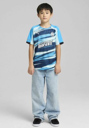 Maglietta da calcio a maniche corte a righe blu e azzurro con logo "Olympique de Marseille", abbinata a jeans azzurri e sneakers nere.
