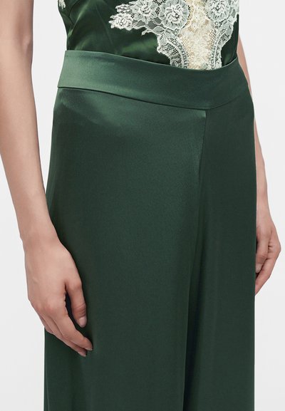 Femme portant un pantalon en satin vert foncé associé à un top vert orné de dentelle, les mains reposent doucement le long du corps.