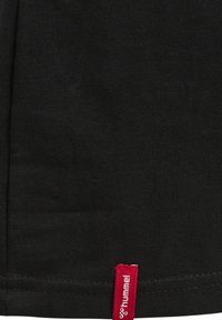 Tissu noir avec une petite étiquette rouge cousue à l'ourlet, affichant le logo et le texte blanc de la marque « hummel ».