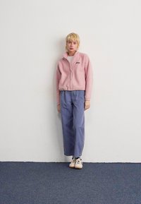 Giovane con capelli biondi corti, che indossa una giacca in pile rosa, pantaloni a gamba larga blu e scarpe da ginnastica bianche, in piedi di fronte a un muro bianco.