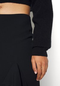 Une personne porte un pull court noir et une jupe noire taille haute avec un détail volanté à l'avant, main détendue le long du corps.