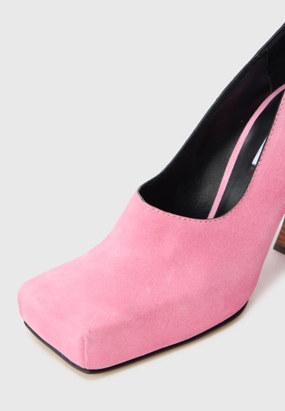 Ροζ σουέτ ψηλοτάκουνο παπούτσι με τετράγωνη μύτη και ξύλινο τακούνι. Το εσωτερικό είναι μαύρο με ορατές ραφές γύρω από το άνοιγμα.