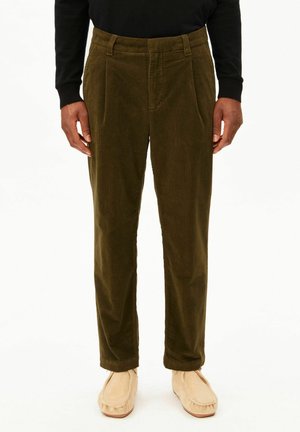 Person trägt olivgrüne Cordhosen, ein schwarzes Langarmshirt und beige Mokassins und steht vor weißem Hintergrund.