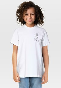 Djevojka s kovrčavom kosom koja nosi bijelu majicu s malom rukom koja pravi gestu srca otisnutom na prsima, stoji nasuprot jednostavne pozadine.