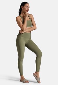 Olivgrüner Sport-BH und hochtaillierte Leggings aus dehnbarem Material, mit einer glatten Textur und minimalistischem Design. Getragen mit beigen Schlappen.