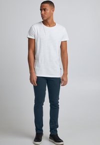 Bela majica z diamantnim izrezom z reliefno teksturo, kombinirana s temno modrimi hlačami ohlapnega kroja in črnimi supergami z belimi podplati.