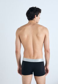 Zwarte aansluitende boxershorts met een zilveren tailleband, gezien vanaf de achterkant, gedragen door een persoon met kort, donker haar tegen een lichte achtergrond.