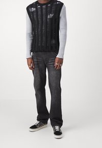 Svart, texturerad stickad topp lagd över en ljusgrå långärmad tröja, kombinerad med mörka jeans och mönstrade sneakers. Silverringar synliga.