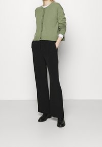 Zelený kardigan s knoflíky, ribbovaným lemem a dlouhými rukávy, zkombinovaný s černými volnými kalhotami a černými kotníkovými botami.