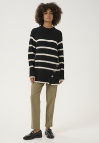 Juoda ir balta dryžuota megzta palaidinė su aukštu apykakle, šoninėmis mygtukų detalėmis ir laisvu siluetu, derinama su pritaikytais chaki kelnėmis.