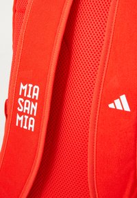 Rød ryggsekk med mesh-materiale, som har hvit tekst "MIA SAN MIA" og en hvit adidas-logo. Teksturert stropp og en livlig farge.