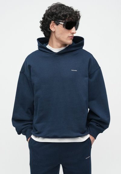 Μπλε σκούρο hoodie με άνετη εφαρμογή, ριμπ μανσέτες και τελείωμα. Διαθέτει μινιμαλιστικό λογότυπο στο στήθος και φοριέται με λευκό T-shirt από κάτω.