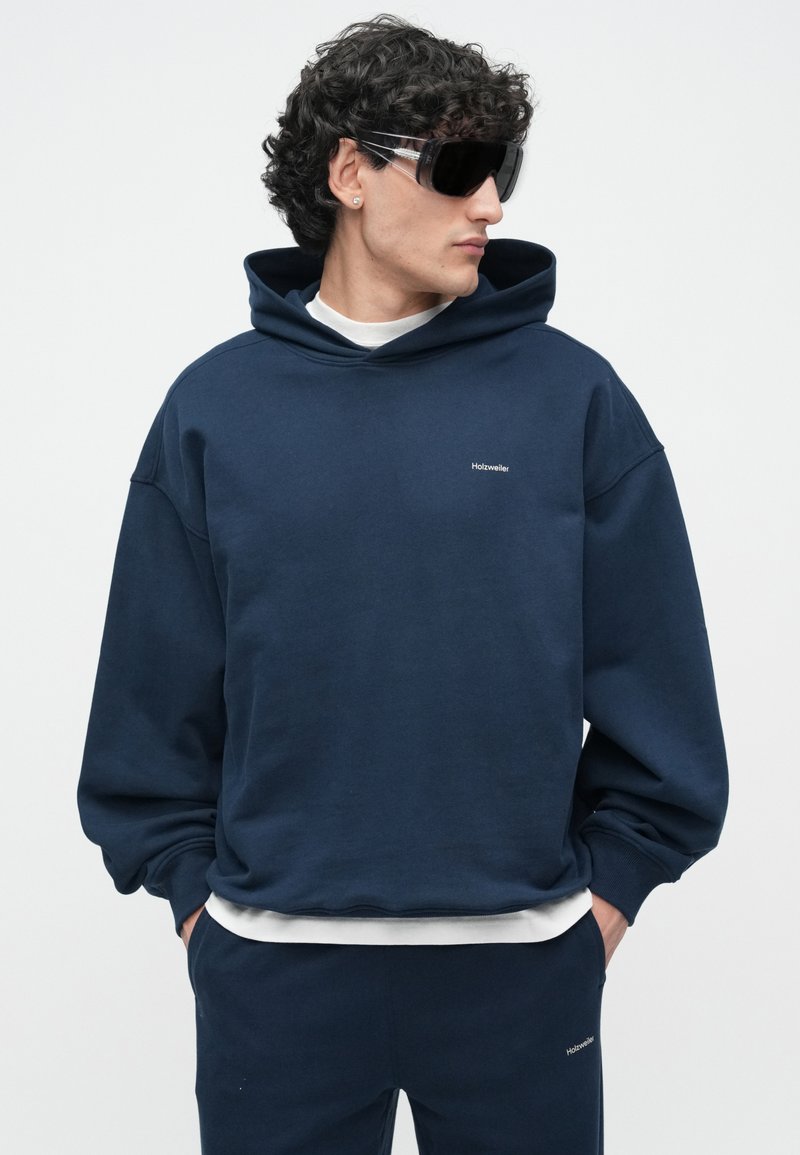 Navy hoodie with a relaxed fit, ribbed cuffs, and hem. Features a minimalist logo on the chest and layered with a white T-shirt underneath.
