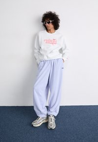 Biely mikina s červeným logom Nike, v kombinácii s svetlo levanduľovými voľnými teplákmi a sivými teniskami. Jednoduché pozadie s modrou podlahou.