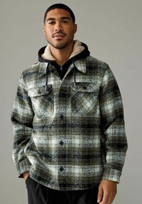 Skridende flanneljakke i nuancer af sort, hvid og olivengrøn med to brystlommer, en sherpa-foret hætte og en knaplukning foran.