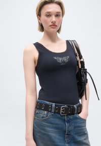 Jeune femme aux cheveux blonds courts portant un haut sans manches noir à imprimé papillon, un jean bleu, une ceinture noire, et un sac à bandoulière noir.