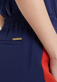 Tela de color azul marino con una cinturilla elástica, que cuenta con un panel lateral rojo y una placa del logo dorado de Michael Kors en el bolsillo.