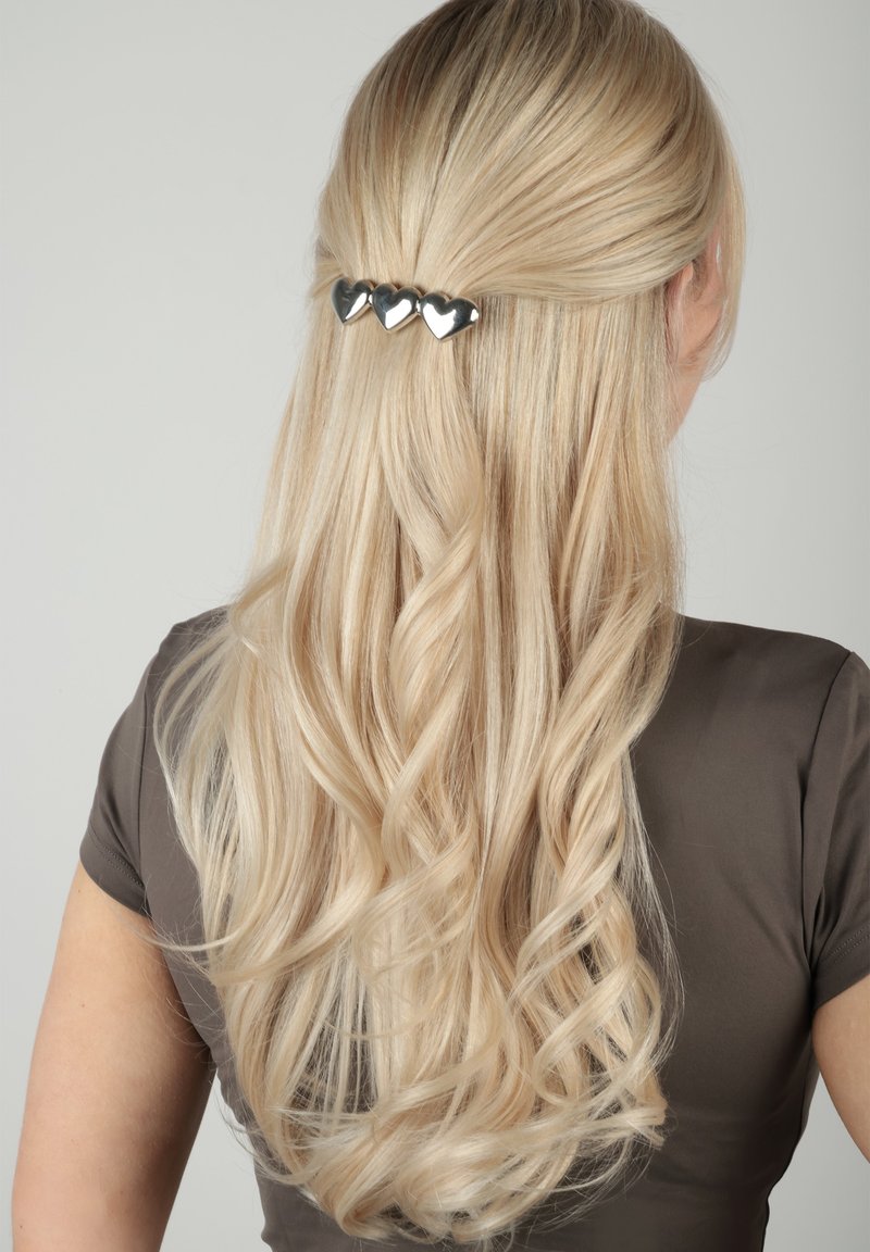 Femme blonde aux longs cheveux bouclés, mi-attachés et maintenus par une barrette argentée ornée de trois formes de cœur, portant un t-shirt marron à manches courtes.