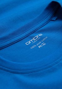 Sininen puuvillapaita, jossa on pyöreä kaula-aukko ja näkyvät saumat. Tunnuksessa lukee 100 % puhdasta puuvillaa ja koko M.