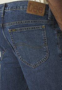 Jeans de mezclilla azul oscuro con una etiqueta de cuero con la marca "Lee", costuras visibles y un clásico diseño de cinco bolsillos.
