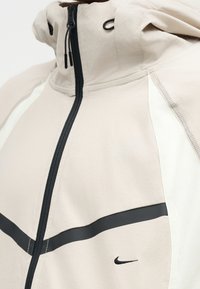 Μπεζ μπουφάν με μαύρο φερμουάρ, μαύρη οριζόντια ρίγα και μικρό μαύρο λογότυπο Nike στο μπροστινό μέρος.