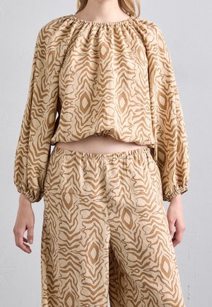 Vrouw draagt beige tweedelig outfit met losse top met lange mouwen en brede broek met abstracte bruine golvende en diamantpatronen.