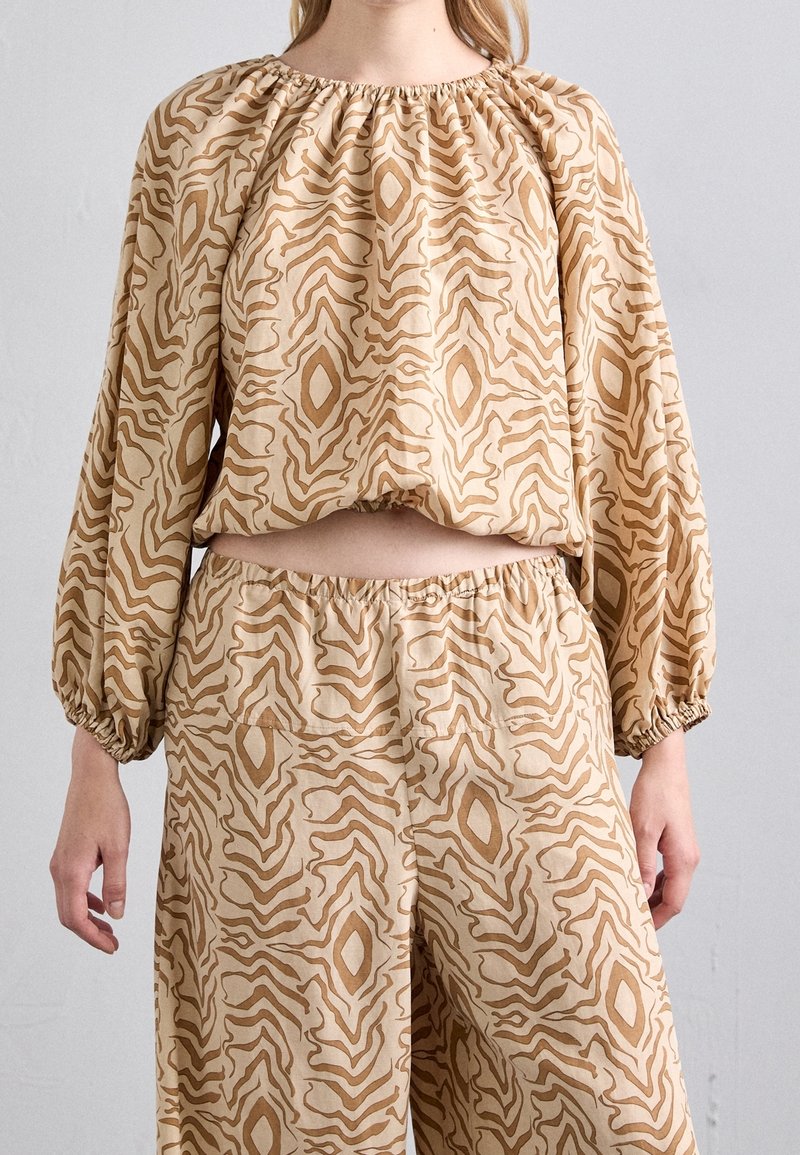 Femme portant une tenue deux pièces beige avec un haut ample à manches longues et un pantalon à jambes larges avec des motifs abstraits marron ondulés et en forme de losange.