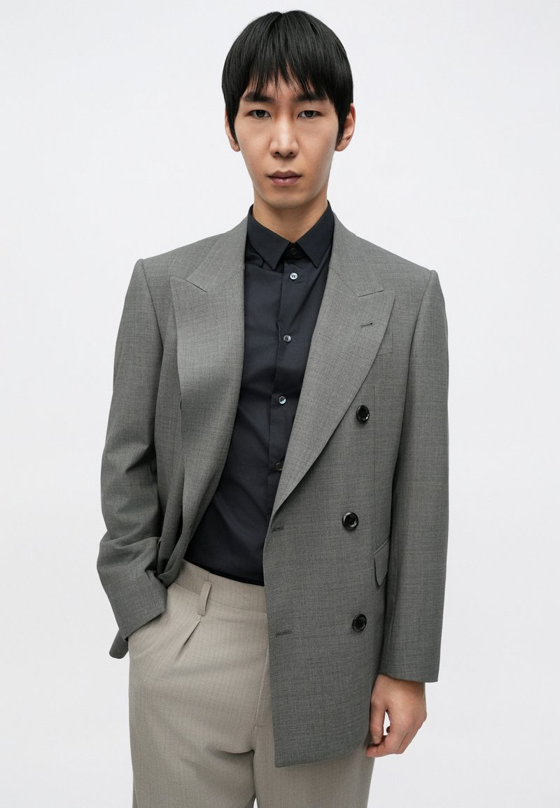 Gray tailored double-breasted blazer with black buttons, paired with a navy shirt. Light beige pleated trousers complete the outfit.