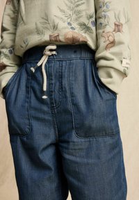 Denim-housut, jotka ovat sinertäviä ja joissa on rento istuvuus, taskut ja nauhakuulostin vyötäröllä. Valaiseva vaaleanvihreä neulepaita täydentää asun.